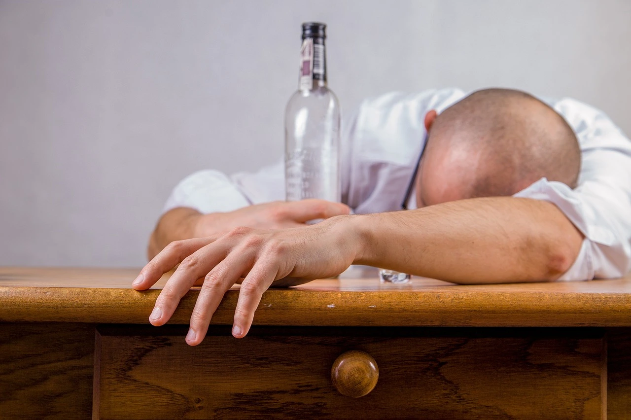 Um homem careca sentado debruçado numa mesa com uma garrafa na mão.