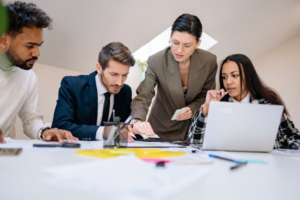 reconhecimento no trabalho 2 homens e 1 mulher sentados na mesa com computador, lupa e caneta e uma mulher em pé ensinando algo
