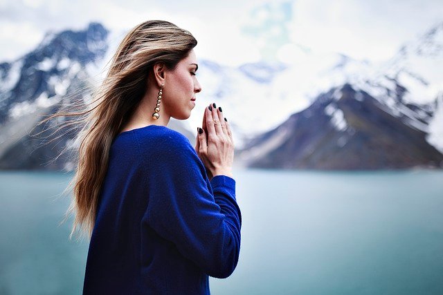 Mulher com blusa azul orando em pé com montanhas de gelo ao fundo da imagem