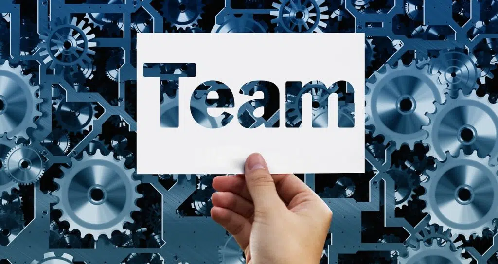 Mudança de Atitude: 50 Frases Incríveis Para Inspirar Sua Equipe uma mão com um papel escrito team em ingles com engrenagens ao fundo