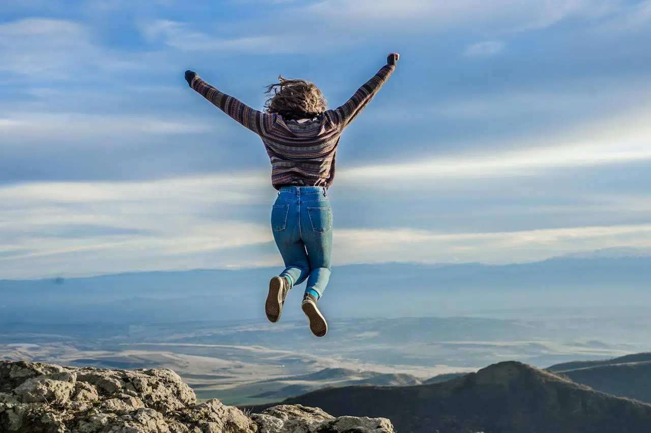 Mudança de Atitude: 50 Frases Incríveis Para Inspirar Sua Equipe Uma moça de braço aberto pra cima pulando em cima de uma montanha