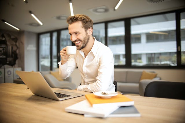 Um homem rindo e sentado segurando uma xícara e olhando para o computador