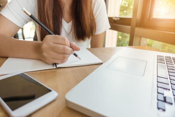 técnica pomodoro garota sentada sem aparecer a cabeça segurando um lápis e escrevendo em um caderno com o celular e computador na mesa